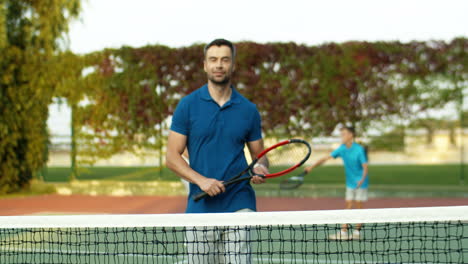 retrato de un hombre guapo que se acerca a la cámara con la raqueta en la mano, se apoya en la red y sonríe a la cámara mientras pasa tiempo con su familia en una cancha de tenis