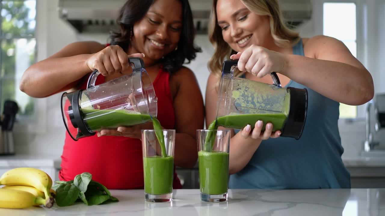 Naklápějící se džbány, dvě ženy nalévající zelené smoothie do vysokých sklenic, sdílející zdravý nápoj.
