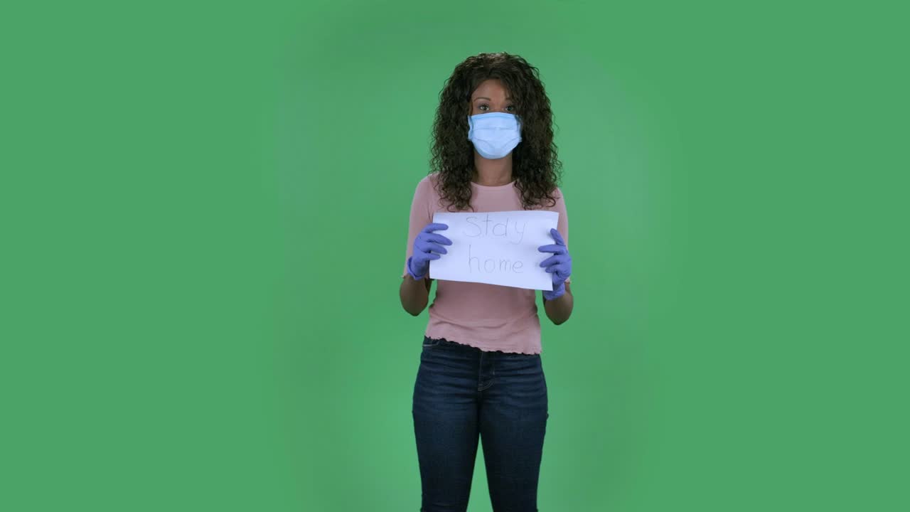 의료 마스크를 입은 아름다운 아프리카계 미국인 젊은 여성의 초상화는 카메라를 바라보고 집에 머물라는 단어가 새겨진 포스터를 들고 있습니다.