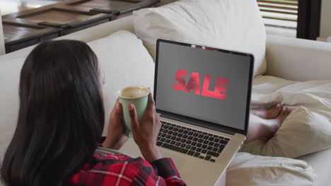 biracial woman on couch with coffee, using laptop for online shopping, slow motion