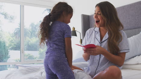 Niña-Hispana-Sorprende-A-Su-Madre-En-El-Dormitorio-Con-Una-Tarjeta-Casera-Para-El-Día-De-La-Madre