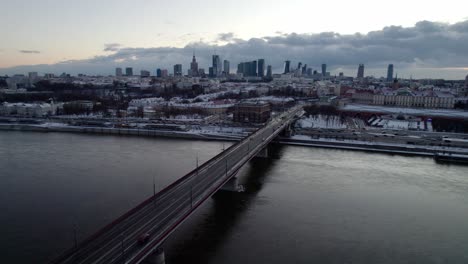 Toma-De-Drones-Que-Muestra-El-Río-Vistula-Y-La-Conducción-De-Automóviles-En-El-Puente-En-Dirección-A-Varsovia-Durante-La-Puesta-De-Sol-En-Invierno---Horizonte-Con-Edificios-De-Gran-Altura-En-Segundo-Plano