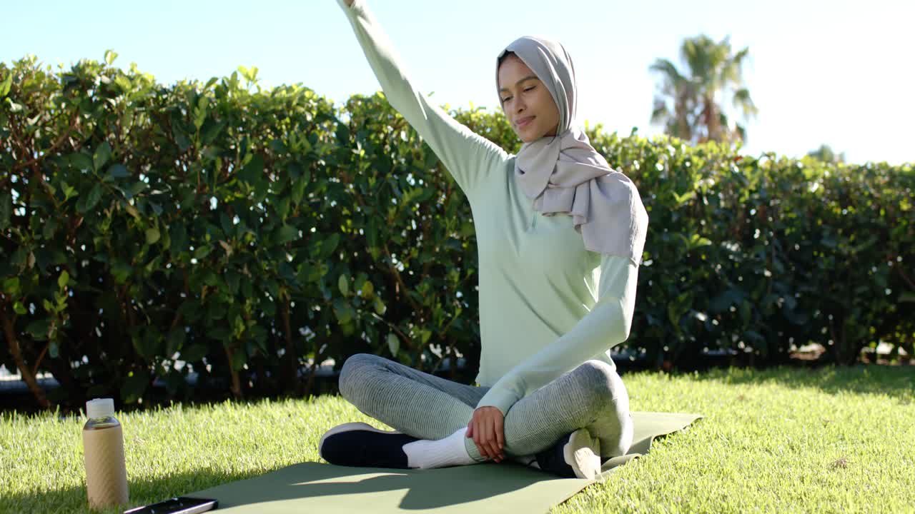 Wanita dewasa memejamkan mata, meregangkan bahu, dan bersantai di matras hijau di halaman rumput, dengan hijab, botol, dan ponsel di dekatnya.
