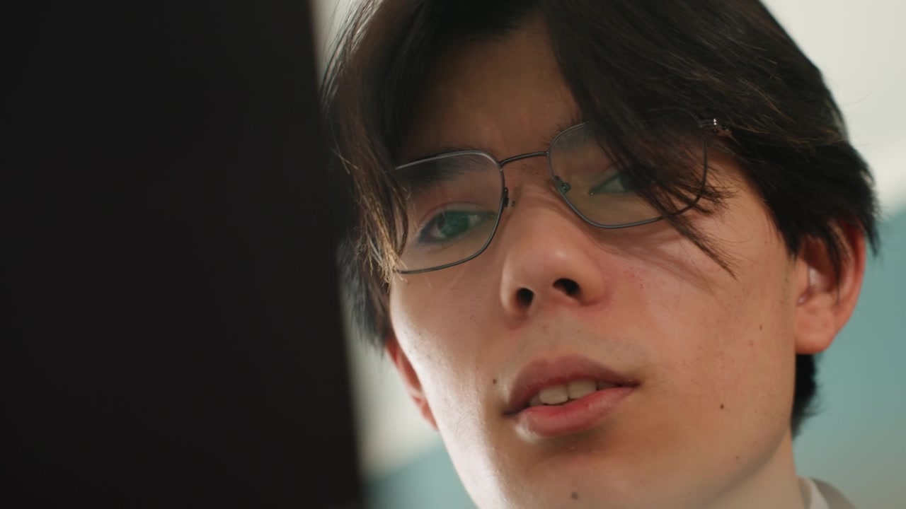ノートパソコンの画面を見るアジア人男性のクローズアップ。眼鏡をかけ、真剣な表情で勉強している。微妙な肌の質感、柔らかな室内照明、ローアングルからの視点。コードに集中する学生のありのままの映像。