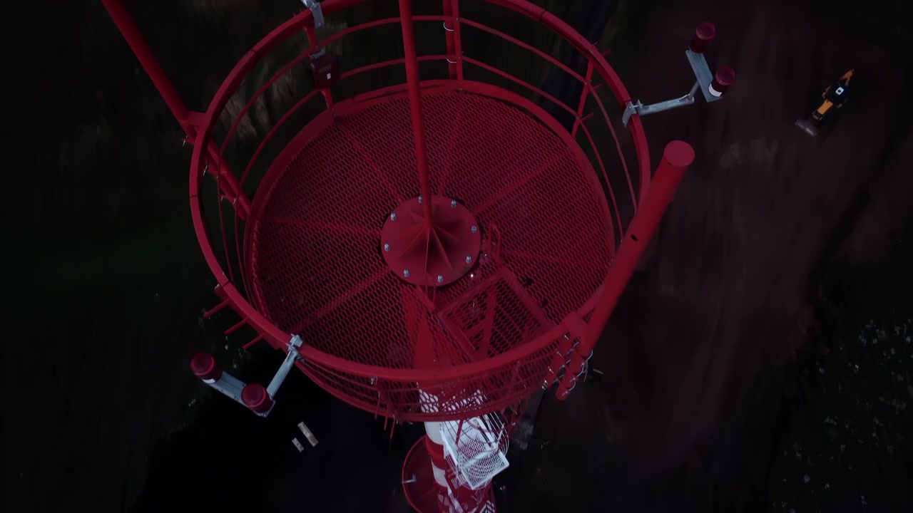 Overhead nighttime view of a red and white industrial tower rising from dark water, featuring a metal ladder, platform, and railings, with a small boat