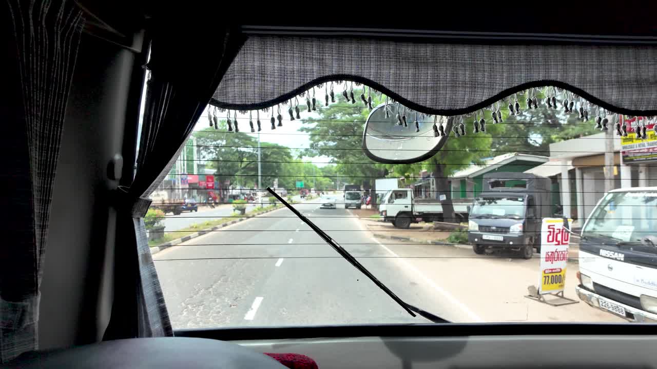 POV perspective from inside a vehicle on a bustling road in Sri Lanka, surrounded by lush greenery and street life.