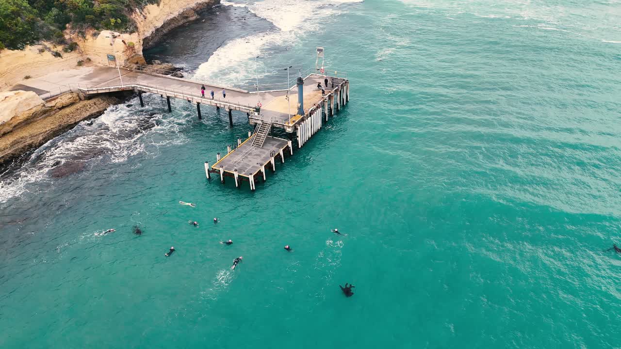 Drone footage captures swimmers near a jetty in Port Campbell, Australia. Clear turquoise waters and coastal landscape create a serene atmosphere