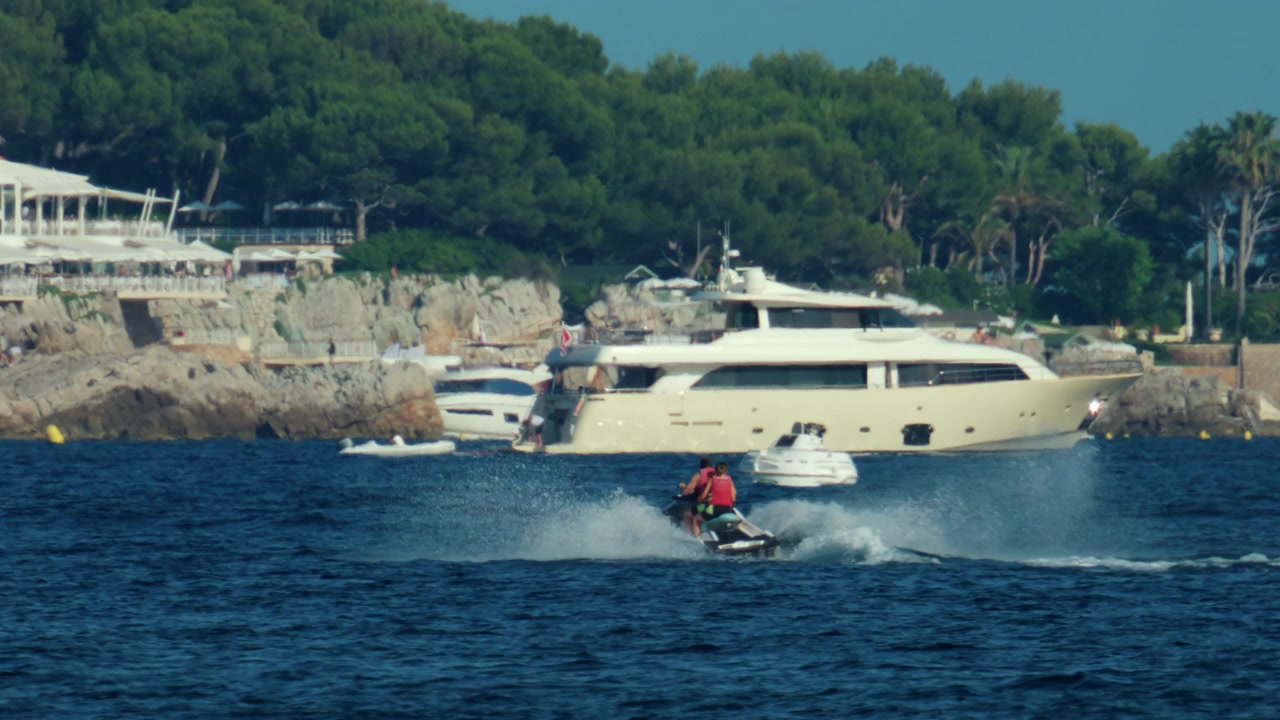 Persone che praticano sport acquatici estivi, sfrecciando su una moto d'acqua e creando spruzzi vicino a una villa costiera.
