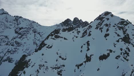 Aerial-steep-snow-icy-mountain-top-slope-flying-towards-snowy-summit-on-sunny-day,-alpine-landscape