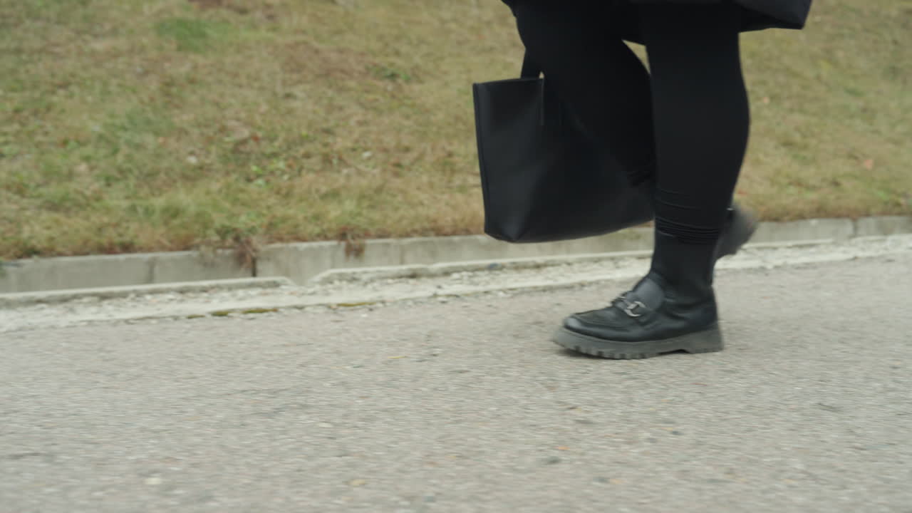 Primo piano ravvicinato dell'asfalto a bassa angolazione, donna che cammina con ritmo da danza, passo svelto e sicuro con una borsa a tracolla, stivali che battono sul marciapiede, dinamico tragitto urbano, andatura e movimento vivaci.