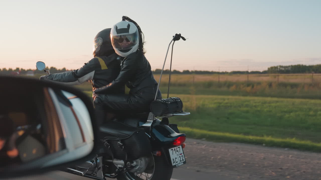Side view of friends wearing helmets riding motorcycle during countryside road trip, passenger embracing rider and looking back while moving through open fields with trees