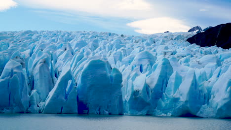 Blue-sunlit-glacier-melting-into-grey-lake,-distant-mountains