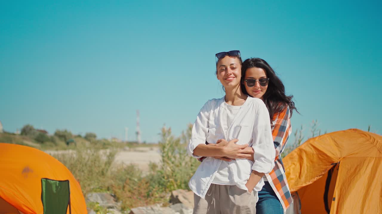 매력적인 아시아 여성이 여자친구에게 와서 뒤에서 그녀를 포옹합니다. 즐거운 소녀는 여름 휴가 동안 무료 캠핑 삶을 즐니다.
