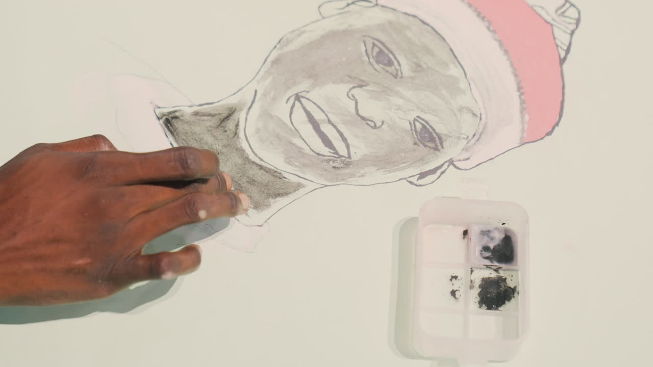 Ritratto di persona di colore dipinto sotto luce soffusa, studente d'arte di colore che traccia e sfuma uno schizzo a carboncino di un soggetto sorridente che indossa un berretto rosa, primo piano su mani dalla pelle scura e vassoio per palette con grafite