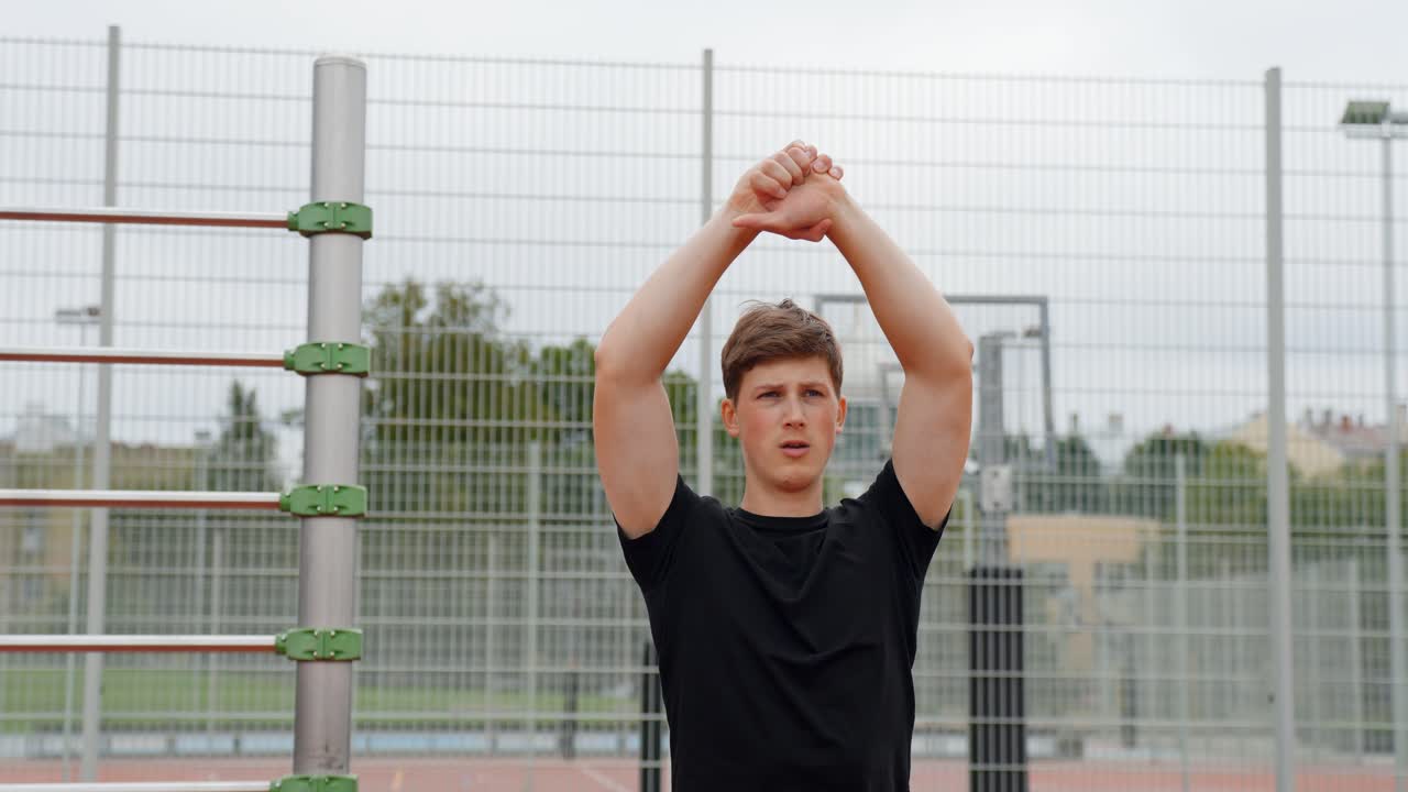 Fixed shot of a young man standing in an outdoor sports area, stretching his back with arms raised above his head