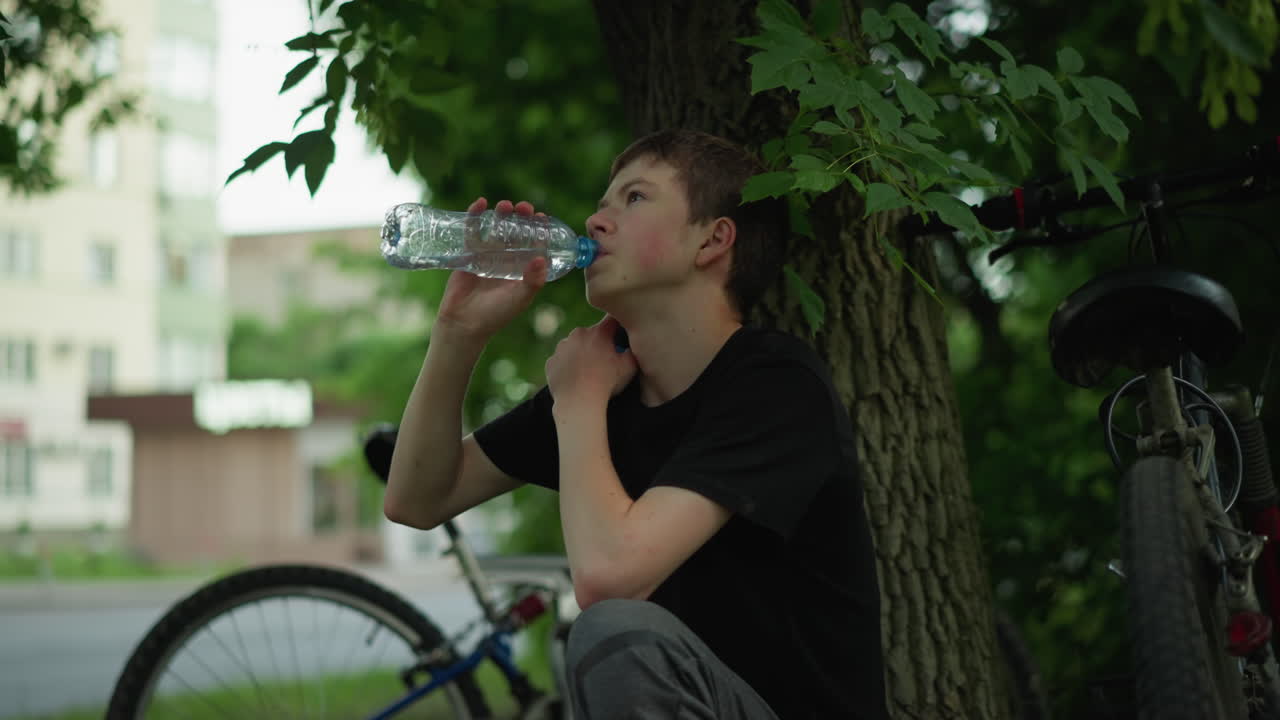 어린 소년은 주차된 자전거 의 나무 에서 휴식을 취하면서 물을 마시며, 거리를 가로지르는 건물의 흐릿한 시야와 함께 차갑게 목을 만집니다.