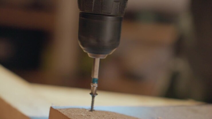 Close up shows drill driving screw through wooden board with steady motion focusing on precision of power tool and material interaction. Drill bit engages wood surface creating clean hole