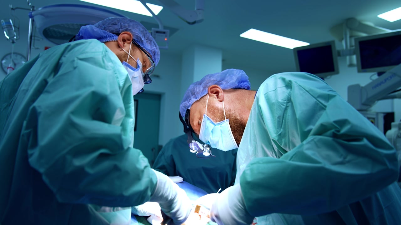 Chirurghi uomini caucasici collaborano a un intervento di chirurgia maxillo-facciale. Il medico, chinato sul paziente, utilizza uno strumento metallico.