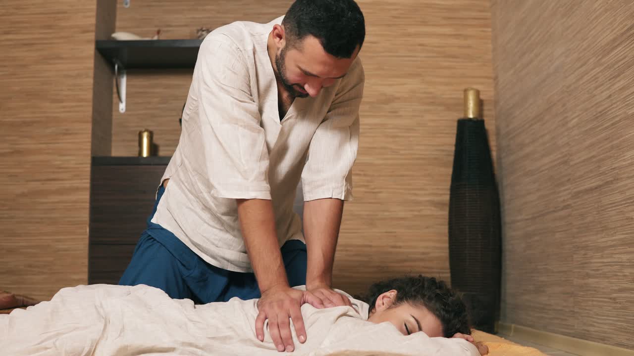 Male thai massagist is treating young woman's back and shoulder. Thai massage session for young attractive woman in kimono. Shot in 4k