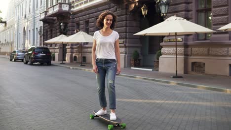 Attractive-brunette-woman-in-white-t-shirt,-blue-jeans-and-white-sneakers-skateboarding-at-sunrise-in-the-city-street-early-in