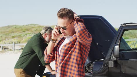 stressed caucasian gay male couple standing by broken down car, talking on smartphone at the beach