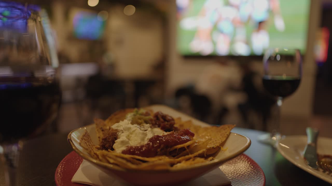 Hand grabs nachos with dip in dimly lit bar, wine glass and sports on television