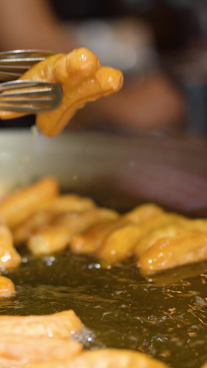 Primo piano di bastoncini di pasta dorati fritti in olio bollente usando pinze metalliche