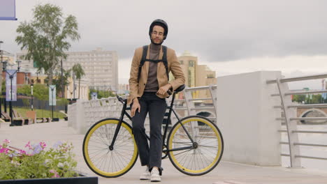 bonito homem afro-americano em roupas formais com capacete e mochila olhando e sorrindo para a câmera enquanto se apoia em uma bicicleta na ponte da cidade