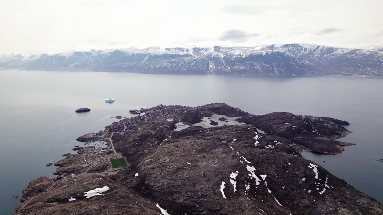 A drone's perspective of the colossal peaks guarding the Uummannaq Fjord and the village Uummannaq. A dramatic flight along the rugged slopes that plunge directly into the cold Arctic water