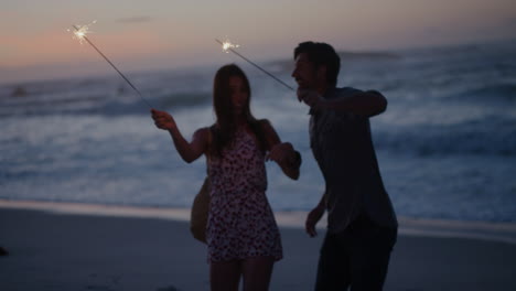 Una-Pareja-Joven-Y-Animada-Ondeando-Bengalas-Bailando-Celebrando-La-Víspera-De-Año-Nuevo-En-Una-Playa-Tranquila,-Fondo-De-Puesta-De-Sol,-Fuegos-Artificiales-Brillantes