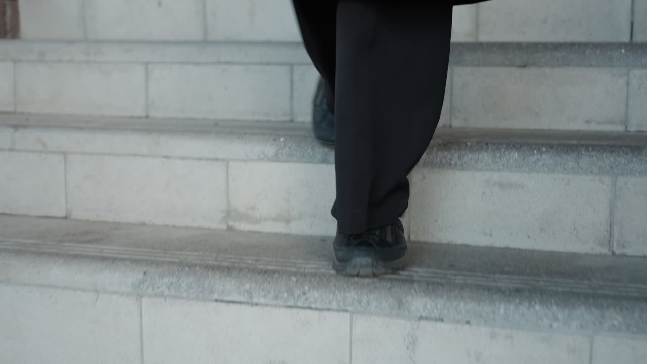 step detail shows person in black shoe moving down concrete stairs, trouser hem brushing riser, texture of worn stone and safety grooves visible with bricks behind, deliberate pace and composition