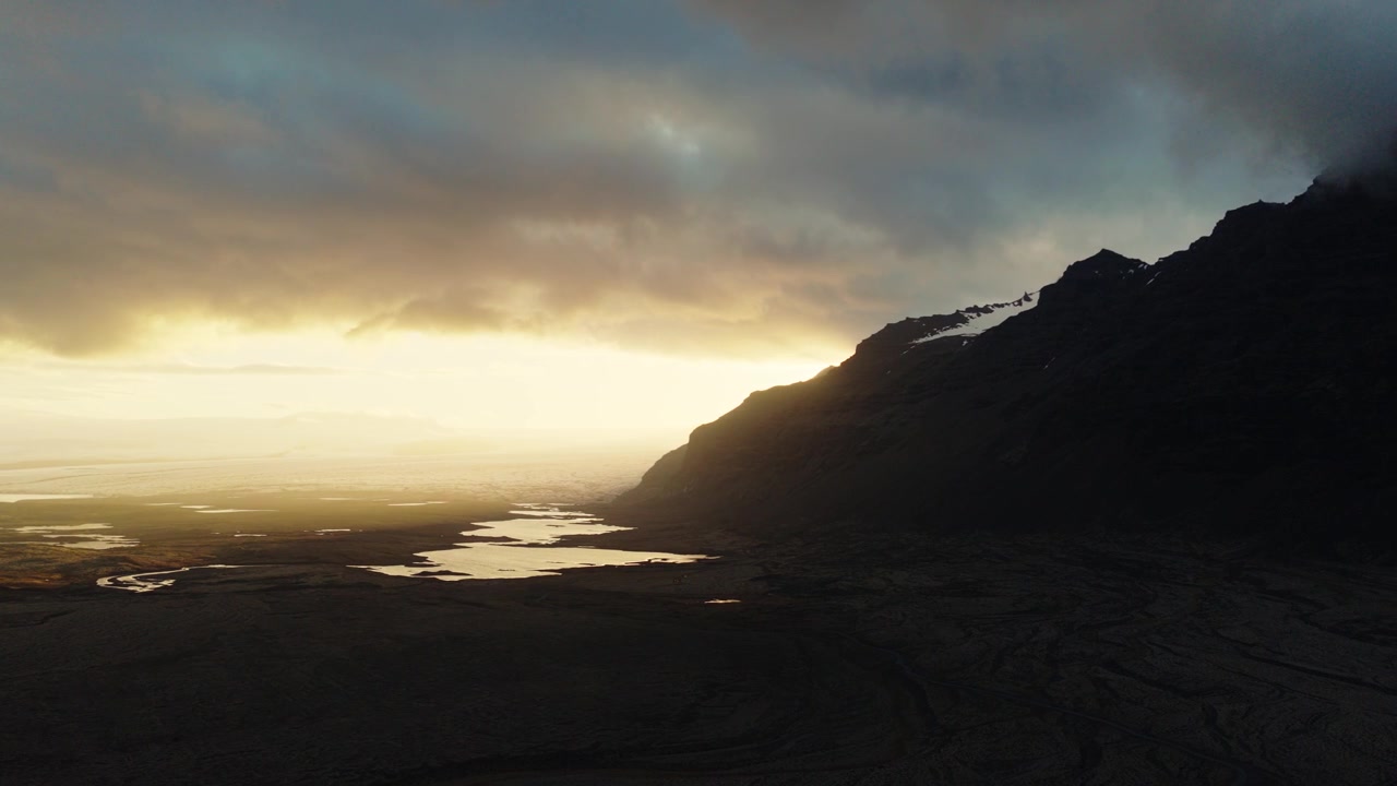 Eyjafjoll mountain slopes rise above Holtsos wetlands in Rangarthing eystra, South Iceland, Iceland, as warm sunset light reaches the coastal lowlands, aerial drone flyover.