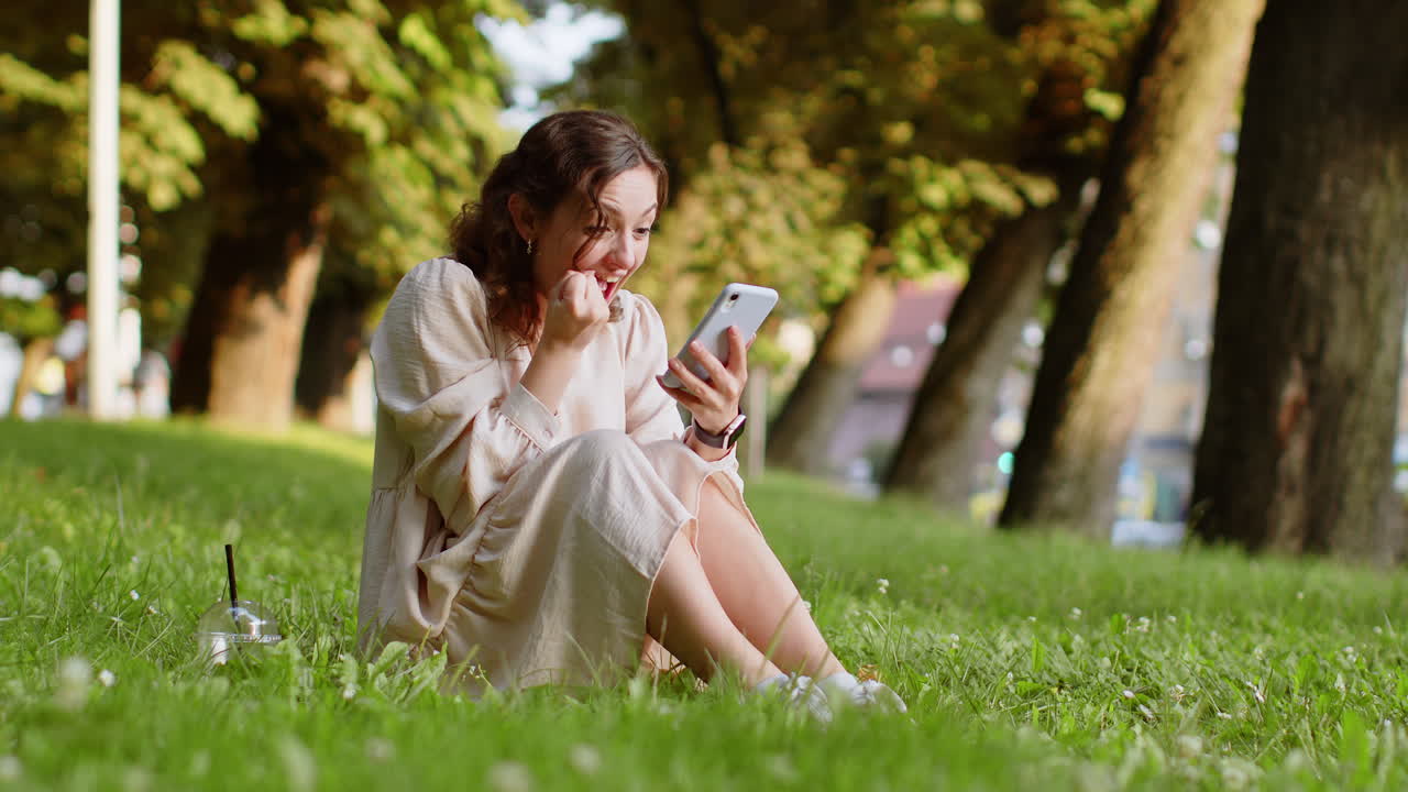 젊은 여성 은 모바일 스마트폰 을 사용 하여 도시 의 공원 밖 에서 좋은 소식 을 축하 하고 있다