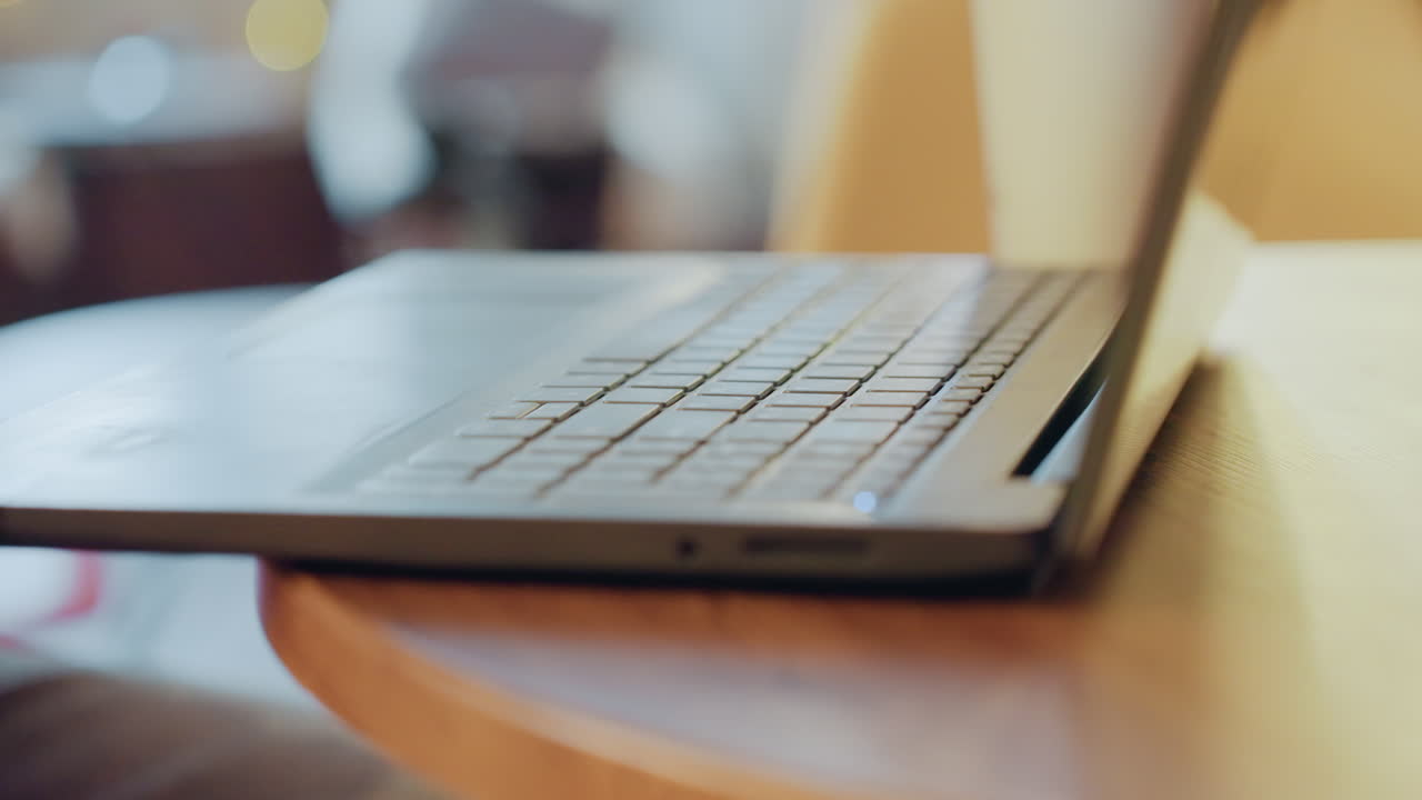 Hvide hænder taster på en bærbar computer ved et hyggeligt cafébord med varm bokeh. Freelance skribent redigerer artikel, studerende researcher noter. Naturligt lys, filmisk nærbillede af tastatur og fingre, bevidst.
