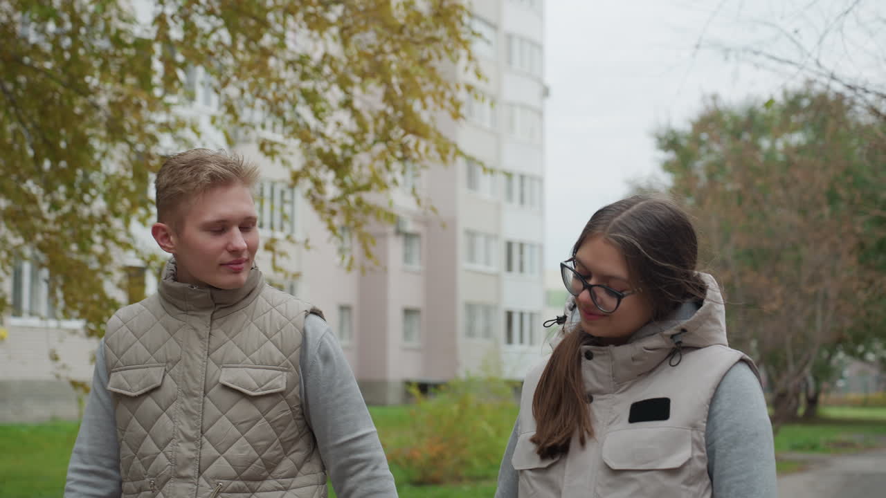 Couple walking side by side outdoors in matching light jackets, holding hands and looking at each other with affection, surrounded by trees and apartment buildings in calm urban environment