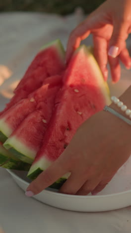 primer plano de la mano de una mujer que alcanza rebanadas frescas y jugosas de sandía en un plato blanco, la luz del sol proyecta sombras suaves, destacando los colores vibrantes de la fruta