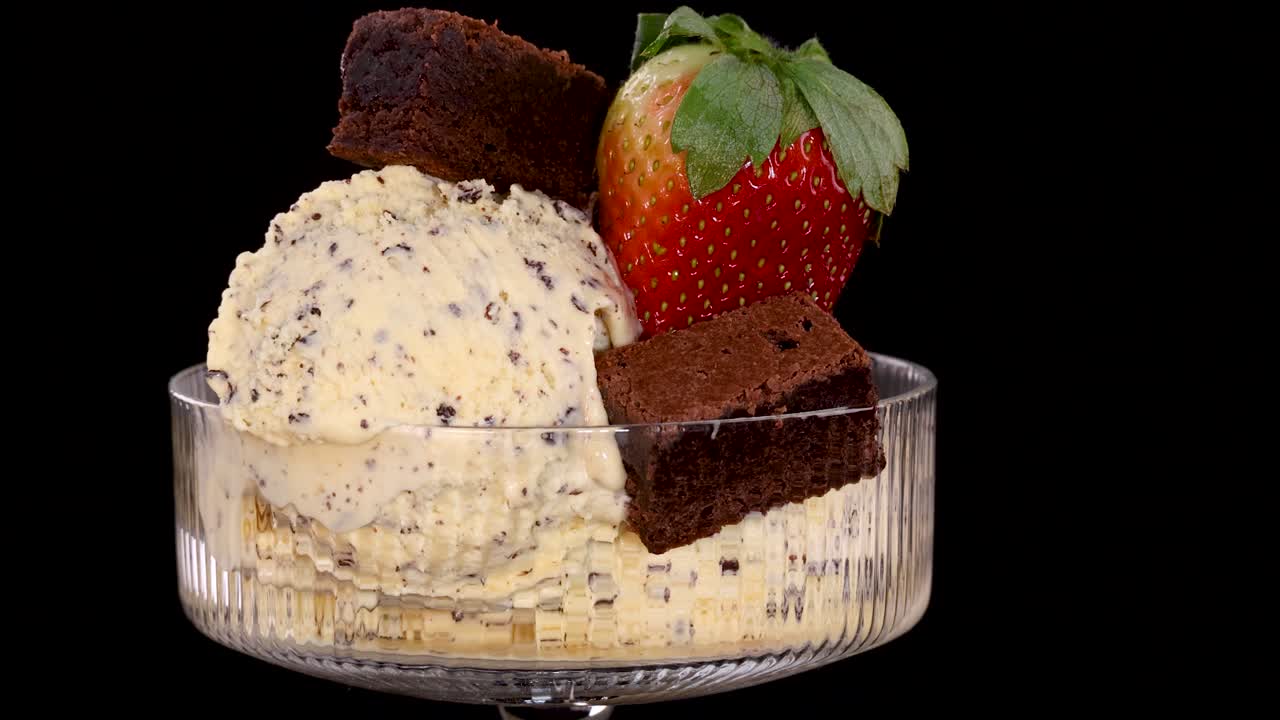 A hand places a fresh strawberry onto a bowl of vanilla ice cream with chocolate chips and brownie pieces, set against a black background with bright, even lighting