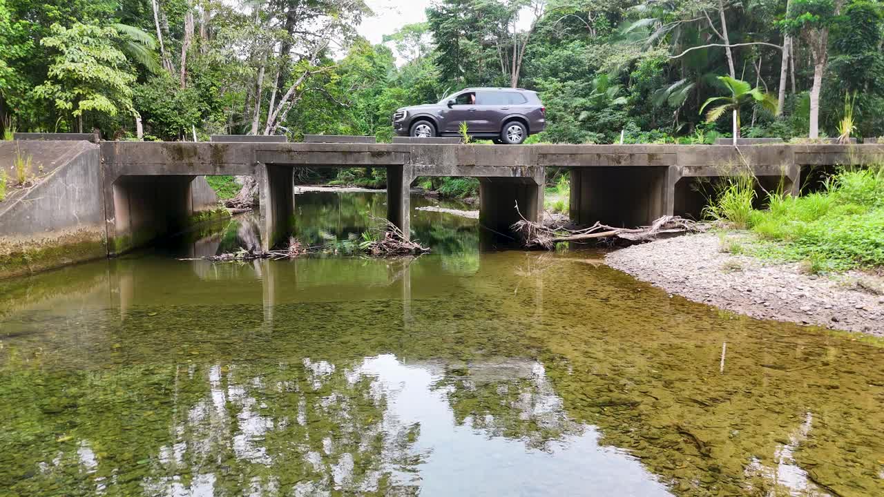 SUV drives across concrete bridge above clear rainforest stream, lush greenery, daylight, steady wide shot