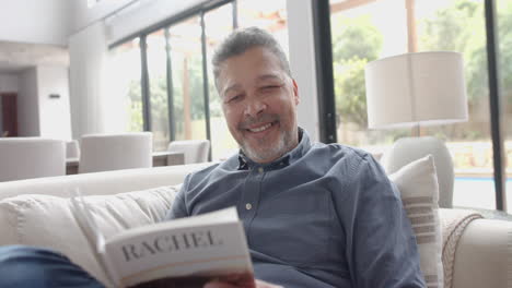 Happy-senior-biracial-man-sitting-on-couch-reading-book-in-living-room-at-home,-slow-motion