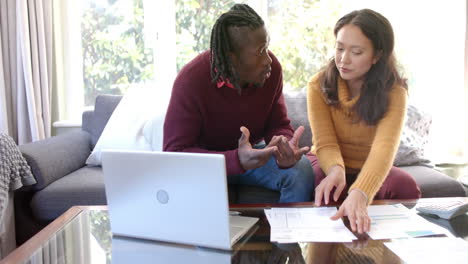 Serious diverse couple with laptop, discussing domestic finances in sunny living room, slow motion
