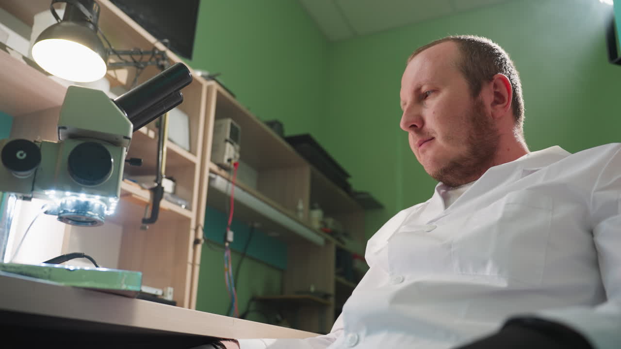 una vista cercana de un técnico en una bata de laboratorio trabajando en un laboratorio bien equipado, el técnico aparece enfocado con una lámpara de escritorio y un estante que contiene algunas herramientas y un microscopio
