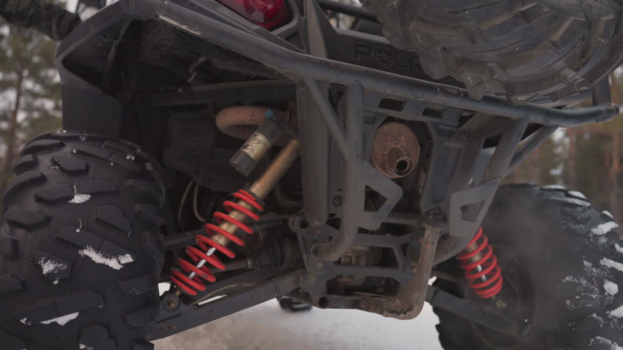 Suspension arrière de VTT dans la neige, amortisseurs à ressort rouges, échappement fumant, gros plan du châssis en contre-plongée, ambiance froide et brute, mécanicien inspectant l'accélérateur et la traction.