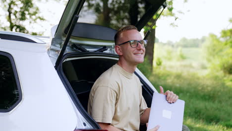 Smiling Caucasian man sits in car trunk. Man has finished work on laptop and is happy about it. Nature at backdrop.
