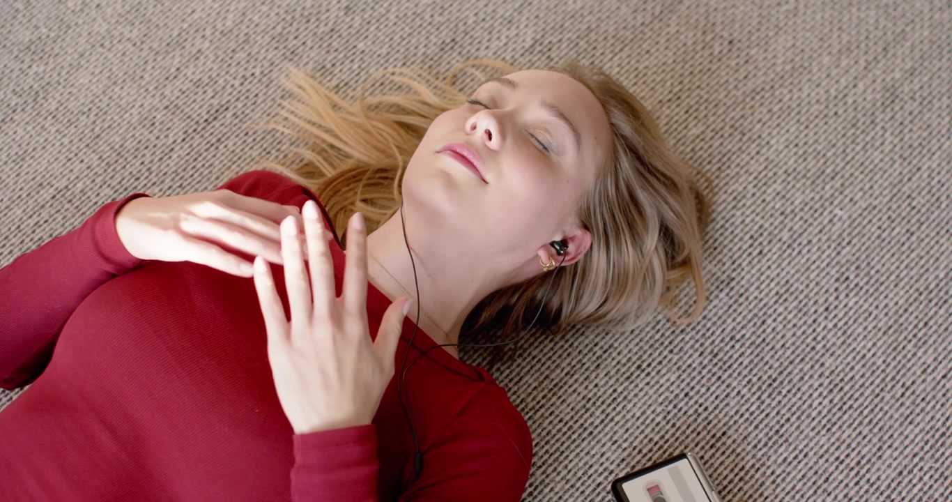 Adult female lying on carpet in red top, wired earbuds pressing cassette, playing music, relaxing