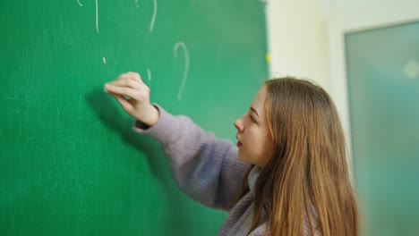 schoolgirl near the chalkboard. girl writing on green blackboard question marks in the classroom. education concept.