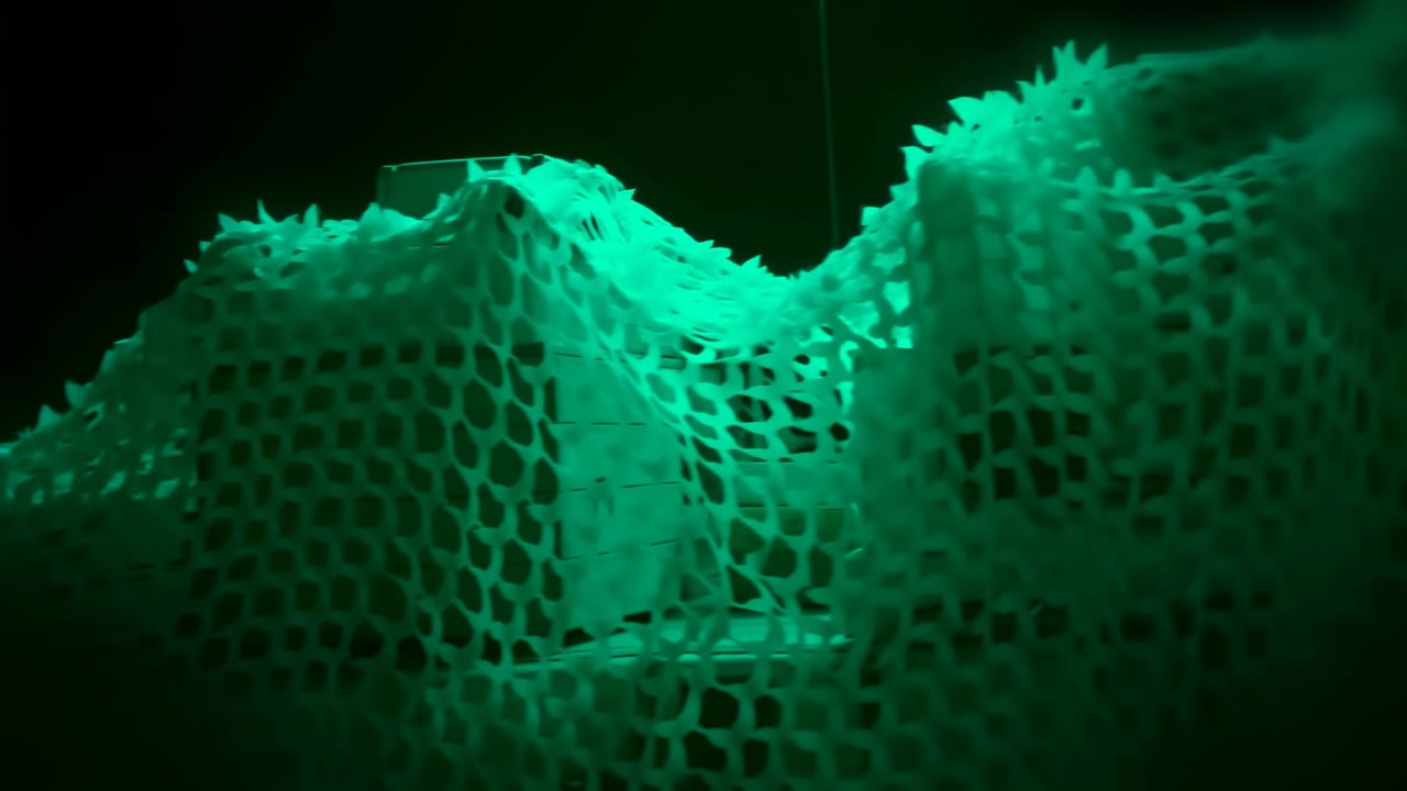Un fotogramma intermedio mostra un tessuto a rete verde luminoso che ondeggia in un ambiente scuro in stile cinematografico.