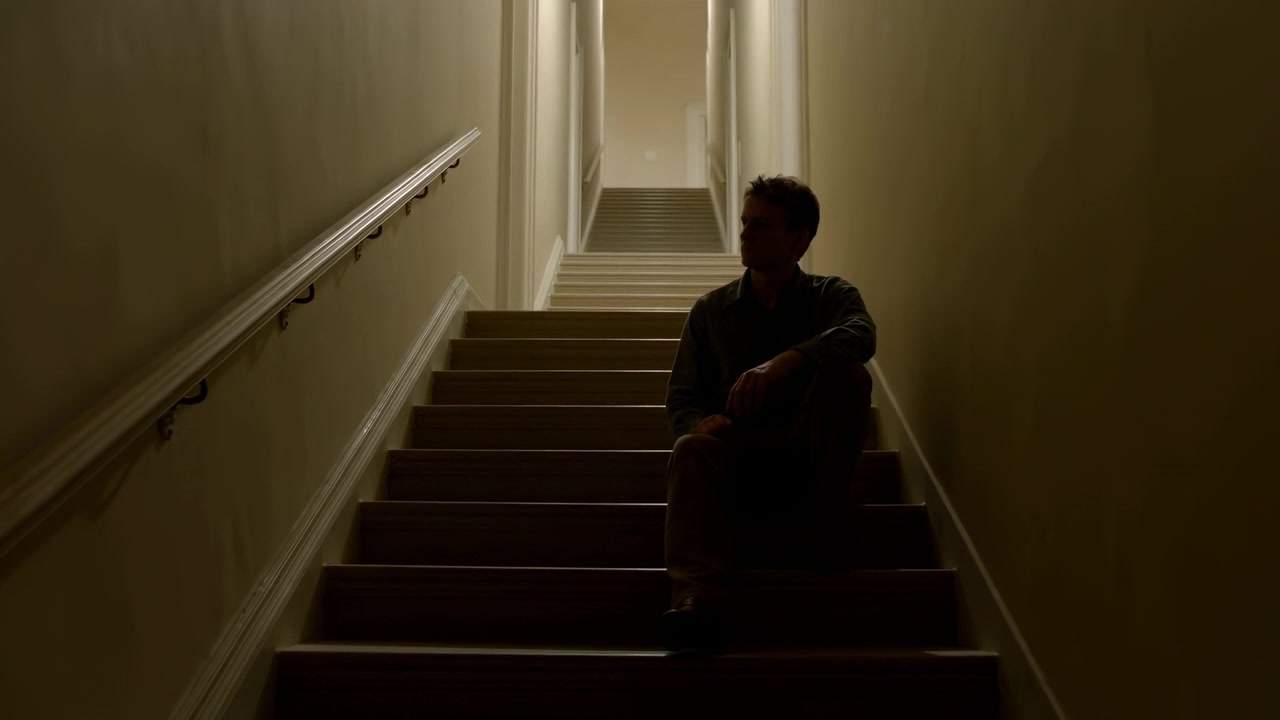 Seated man stays on lower steps while framing gradually widens to reveal more of narrow stairwell, handrail, beige walls, dark carpet, and softly illuminated landing above.