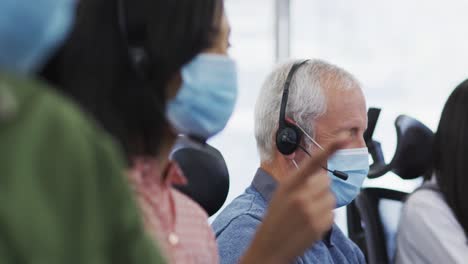 Personas-Que-Usan-Mascarilla-Y-Auriculares-Trabajando-Mientras-Están-Sentadas-En-Su-Escritorio-En-La-Oficina