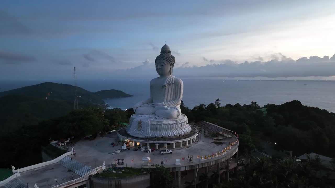 Big Buddha statue in meditative pose on hilltop plaza overlooking ocean, surrounded by trees and visitors, captured under soft cloudy sunrise or sunset sky in Phuket, Thailand