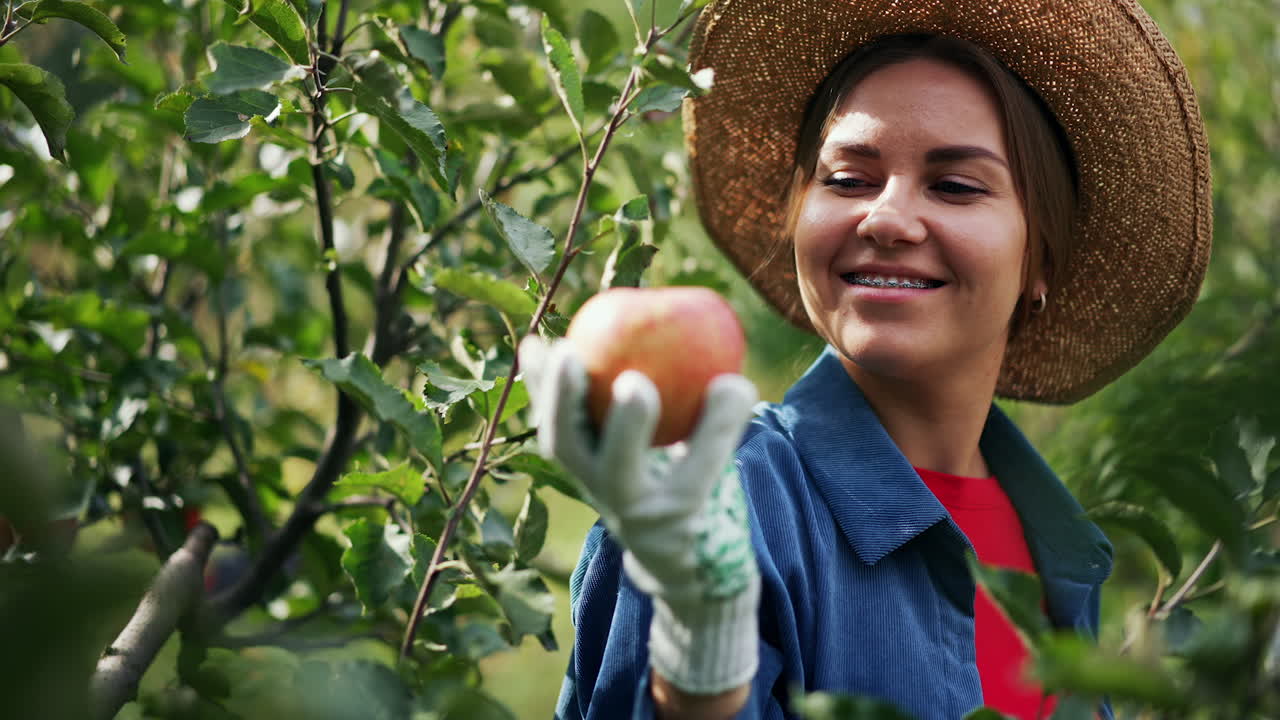 Smiling happy lady in hat enjoys the harvest of apples she picks. Young female farmer is satisfied with the crop of fruit in her garden.